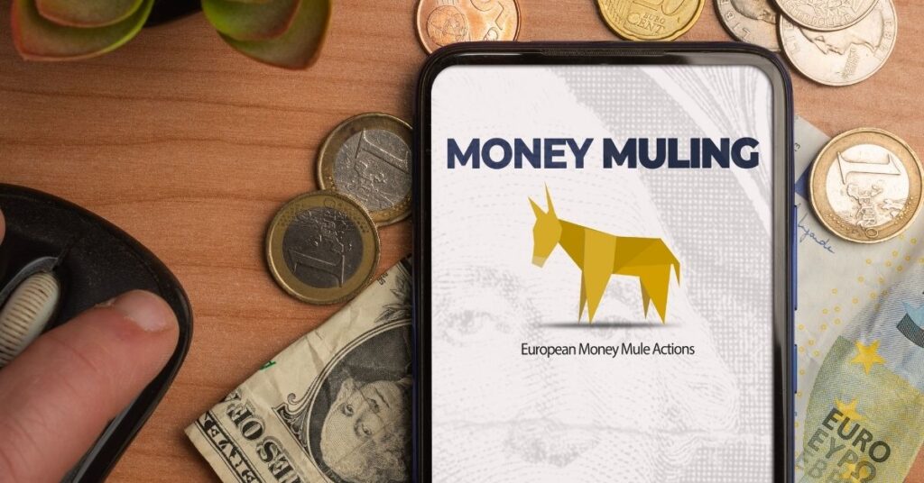 The words "Money Muling" on a mobile phone screen, surrounded by coins and notes on a wooden desk.