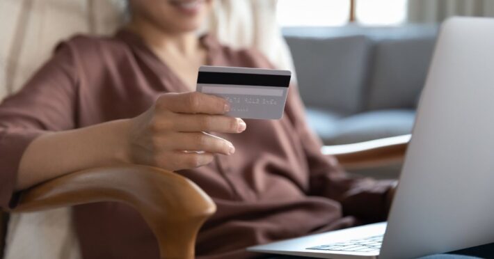 A woman looks at her credit card while working at her laptop.