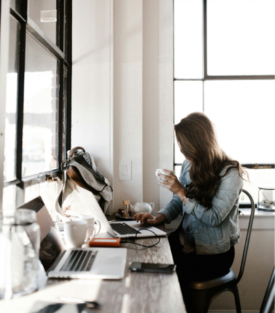A woman sits at a desk with her laptop, drinking from a cup.