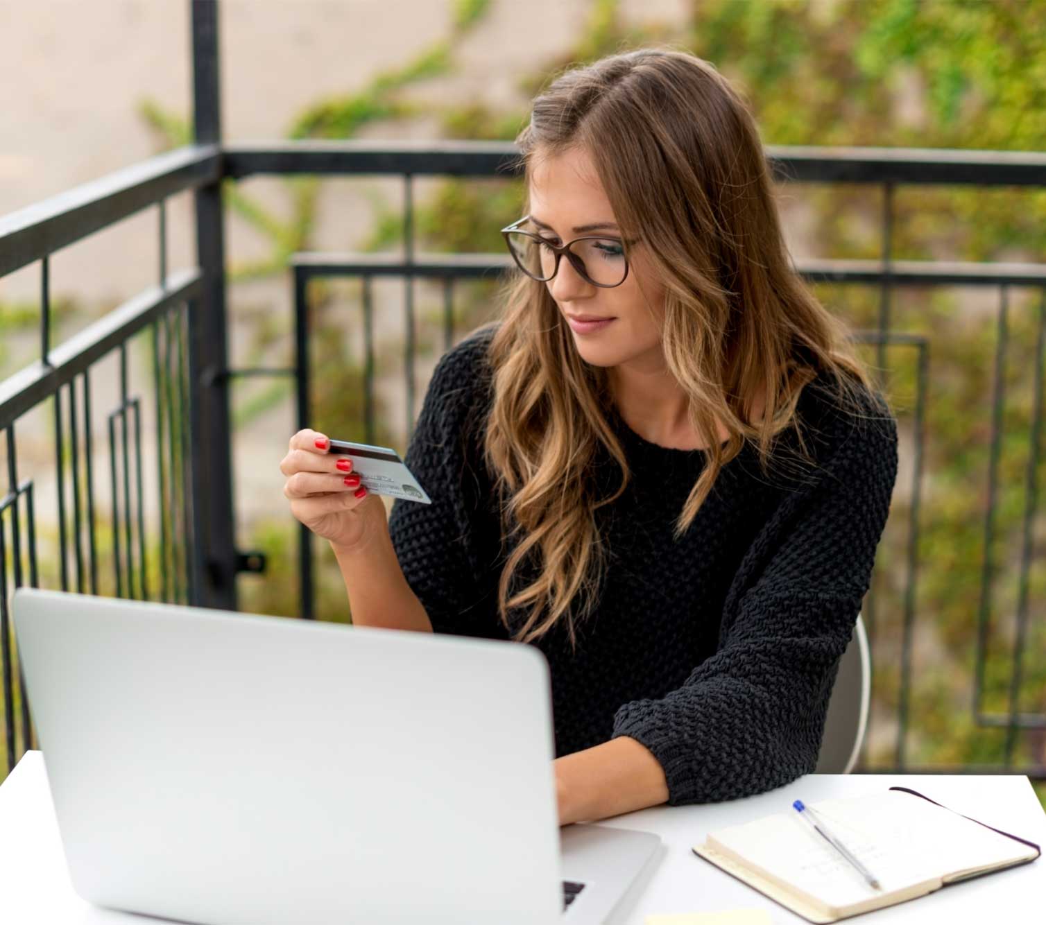 A woman looks at her credit card while typing on a laptop.