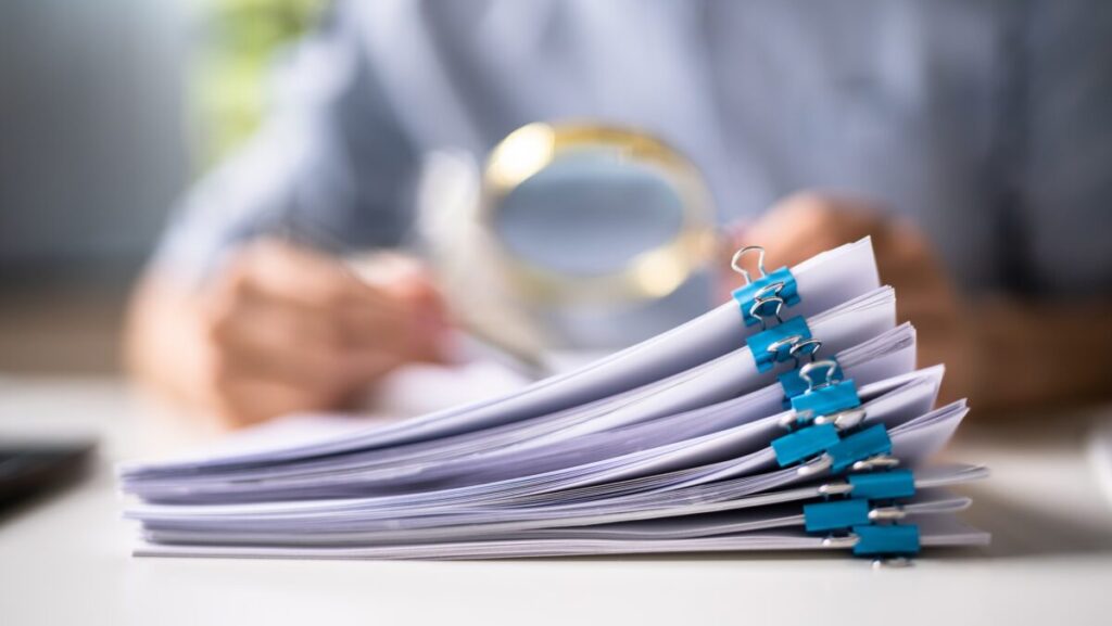 A person investigates a stack of papers with a magnifying glass.